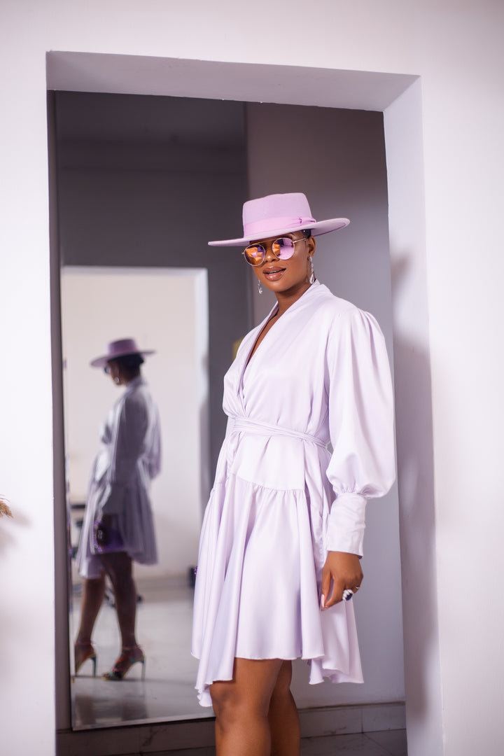 A woman posing in an unlined white silk dress. She is pictured facing to the left and is looking at the camera. She is styled with a purple hat, a purple purse, and silver jewelry.